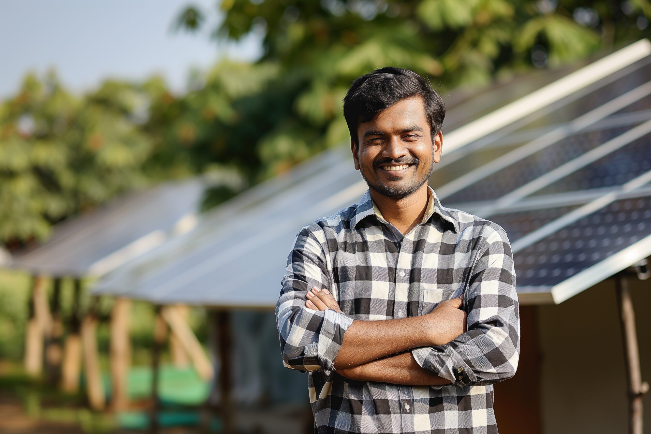 Indian man poses confidently in front of solar panel array with grid-like pattern. Wears black, white checkered shirt, crossed arms. Green background with trees bushes creates natural setting.